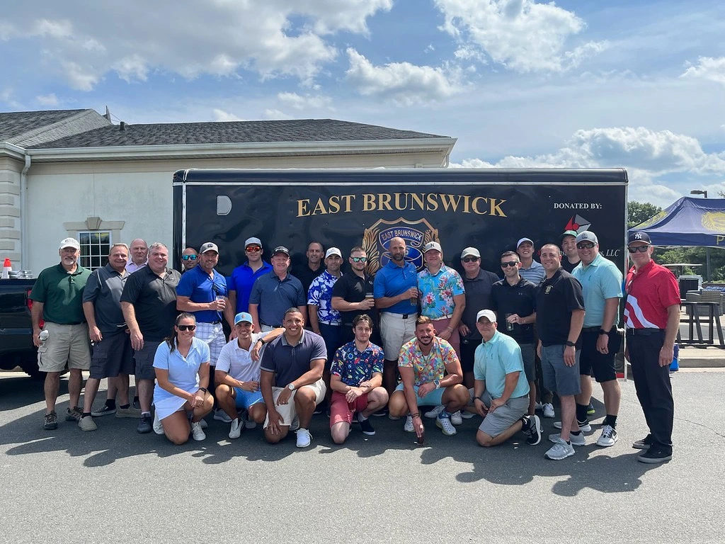A large group of men and women posing together for a photo at a golf event, standing in front of a trailer marked 'East Brunswick.' Some wear golf attire and casual shirts. The background includes a partly cloudy sky.