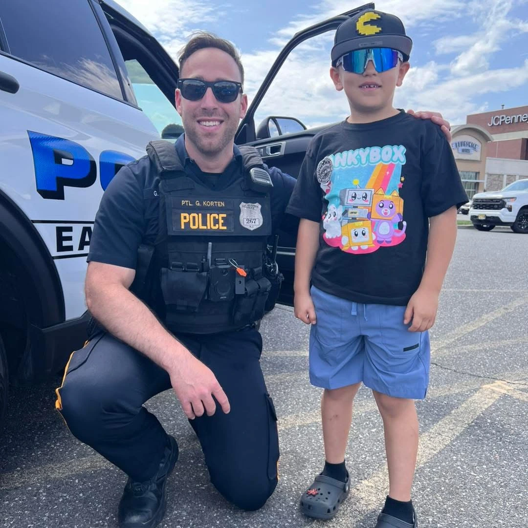A police officer kneels beside a young child wearing a colorful t-shirt and shorts, with a police vehicle visible in the background.