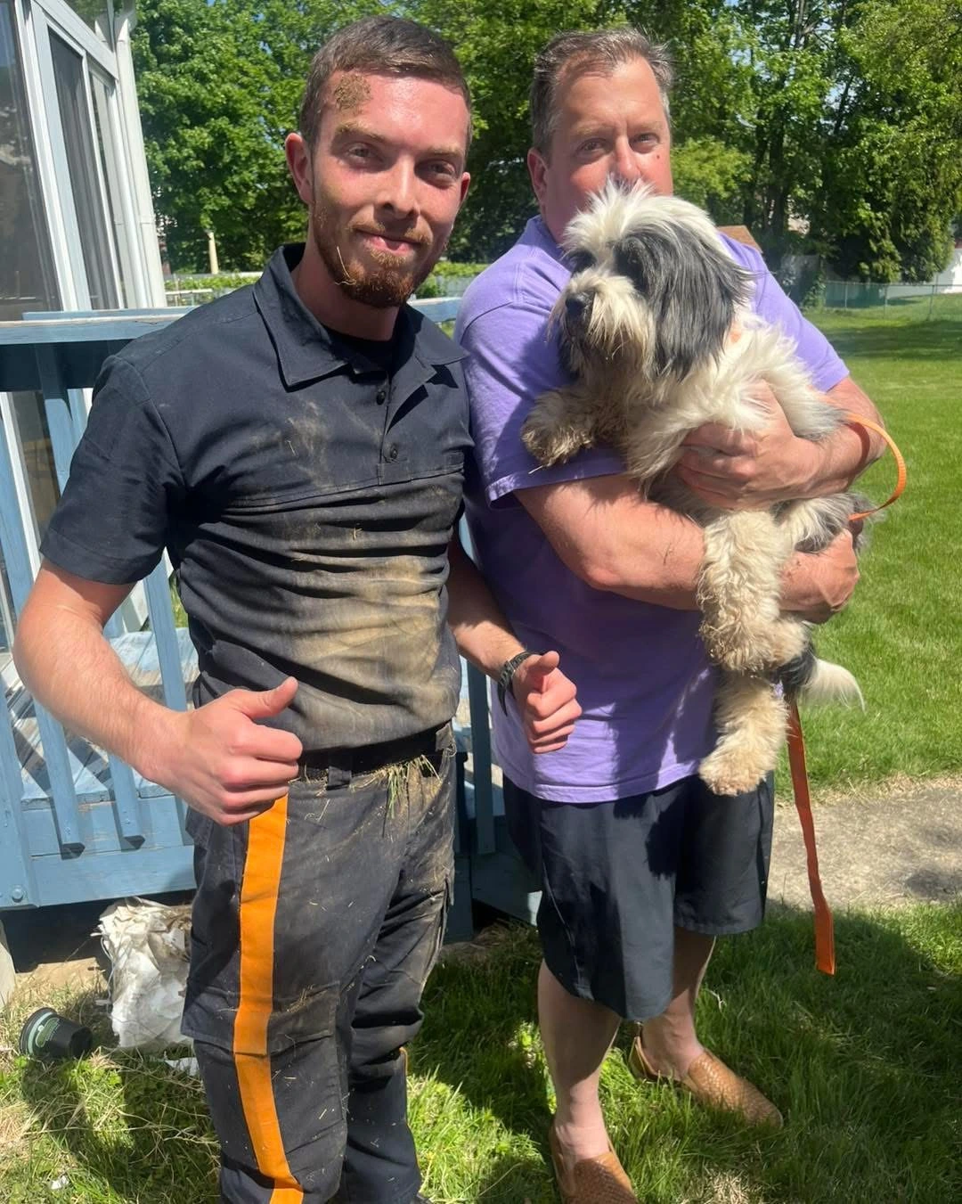 Two men pose together outdoors, one dirty from work, holding a fluffy dog, with greenery and a deck in the background.