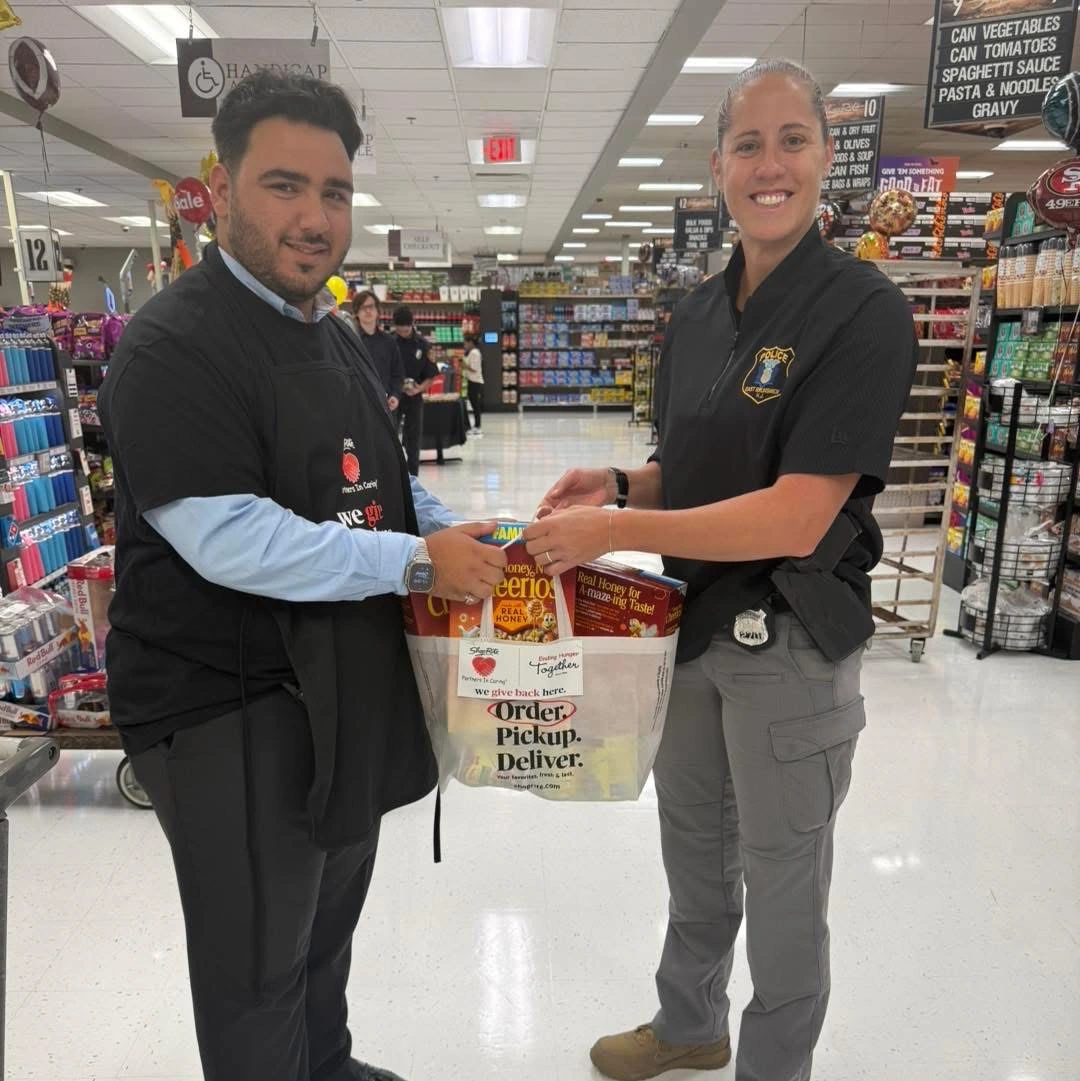 Two individuals are exchanging a bag filled with groceries in a grocery store aisle, surrounded by various products on shelves.