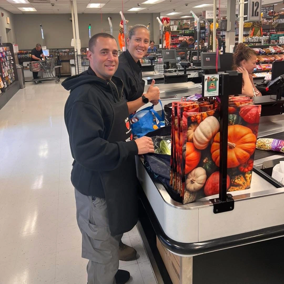 A man in a black hoodie and apron stands at a grocery checkout, bagging items next to colorful reusable bags featuring pumpkins.