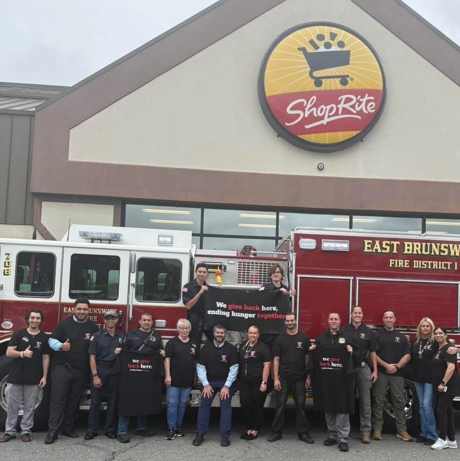 A group of community members and firefighters pose in front of a ShopRite, holding a banner promoting hunger relief initiatives.