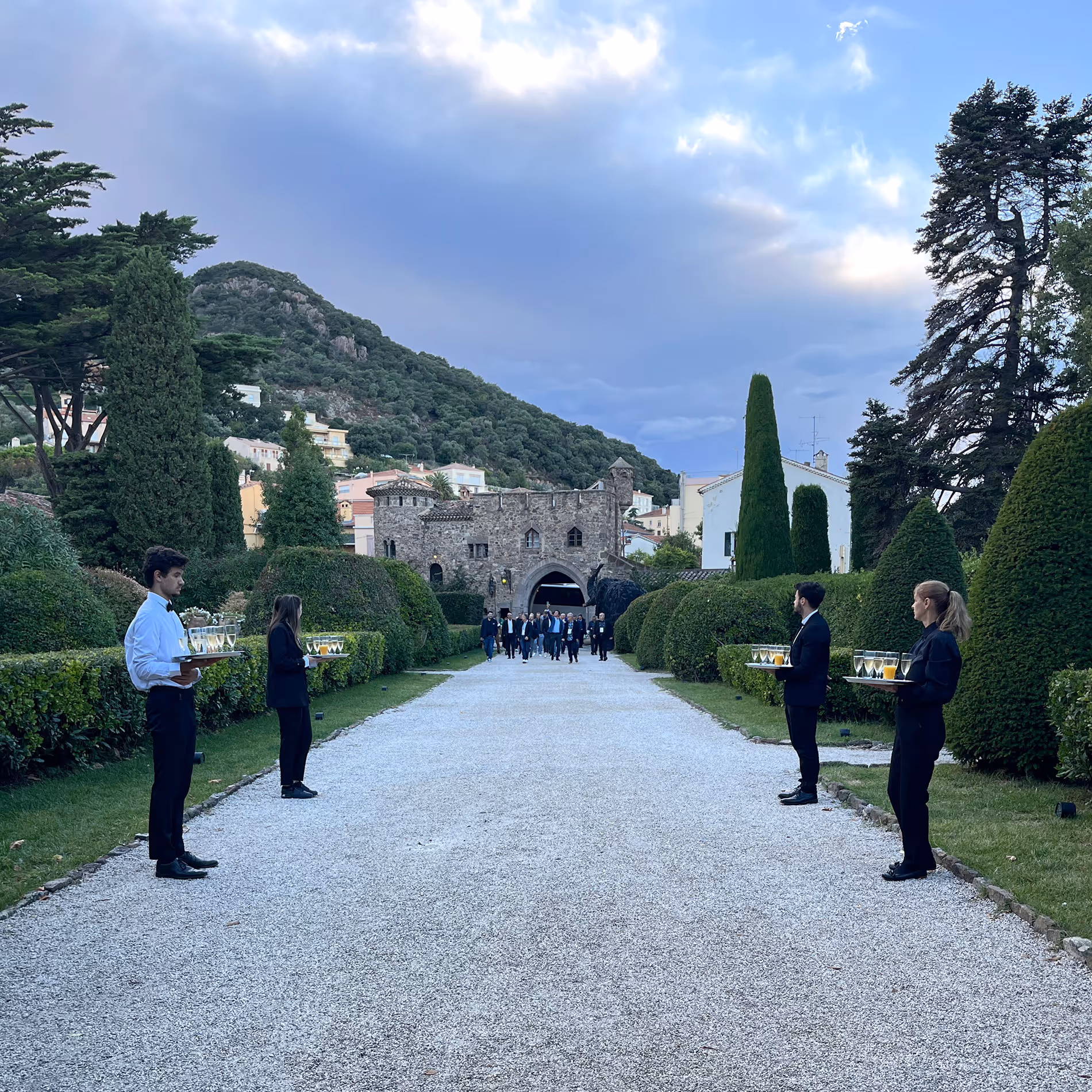 Entrée d'un domaine avec au loin un château et sur les côté des serveurs et serveuses.