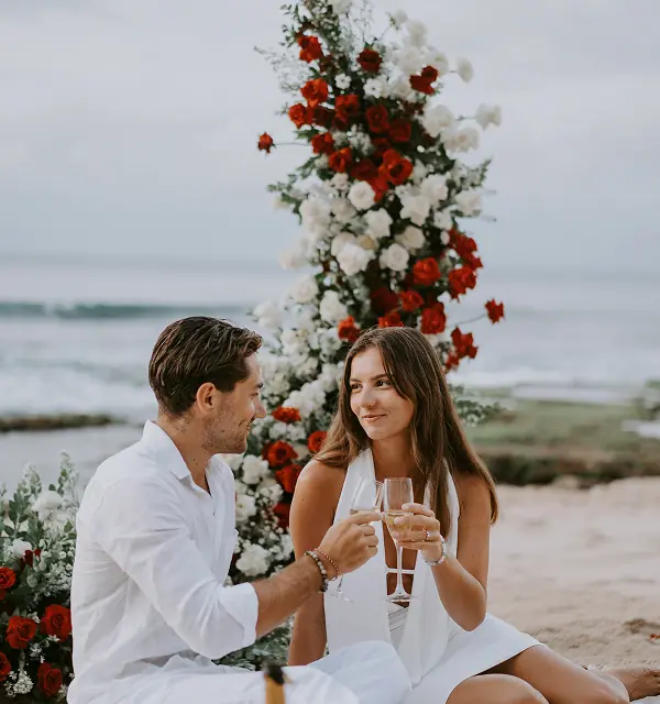 Toast romantique d’un couple devant une arche de fleurs sur la plage au coucher de soleil à Bali, après une demande en mariage.
