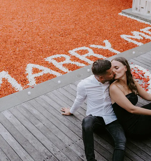 Scène romantique de demande en mariage à Bali, couple français près d’une piscine recouverte de pétales de fleurs rouges avec "Marry Me" écrit en pétales de fleurs blanches.