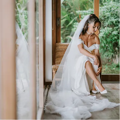 Mariée assise près d'une baie vitrée en train de se chausser, portant une robe blanche et un long voile, entourée de végétation tropicale à Bali.