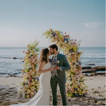 Couple de mariés français qui s'embrassent sur la plage à Bali, devant une arche de fleurs colorées avec l'océan en arrière-plan.