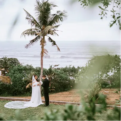 Un couple en tenue de mariage échangeant leurs vœux sous un palmier avec vue sur l'océan à Bali.