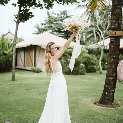 Jeune mariée souriante en robe blanche tenant un bouquet de fleurs levé dans un jardin tropical à Bali.