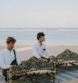 Deux musiciens balinais en tenue blanche jouent d'un instrument traditionnel sur une plage de sable blanc à Bali.