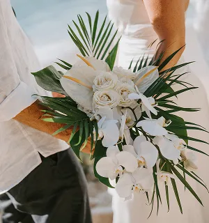 Un bouquet de mariage blanc avec des roses, des orchidées et des feuilles de palmier tenu par un couple français en tenue blanche.