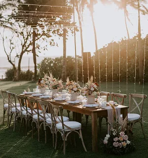 Table de banquet en bois avec décorations florales et vaisselle, éclairée par des guirlandes lumineuses en extérieur au coucher du soleil avec des palmiers en arrière-plan, lors d'un mariage sur-mesure à Bali.