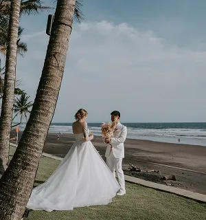 Couple de mariés habillés en blanc près de palmiers sur une plage au bord de l'océan à Bali, avec des palmiers autour.