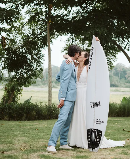Couple en tenue de mariage bleu et blanc posant avec planche de surf "break" dans un cadre tropical à Bali.