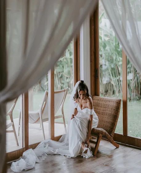 Mariée en robe fluide blanche préparant ses chaussures dans une villa ouverte à Bali, vue jardin.