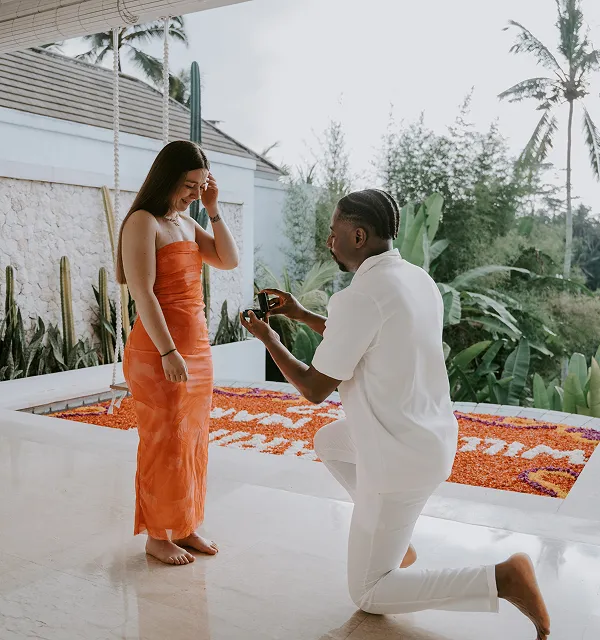 Homme à genoux en tenue blanche faisant une demande en mariage avec une bague à une femme en robe orange, dans une villa avec jardin tropical.