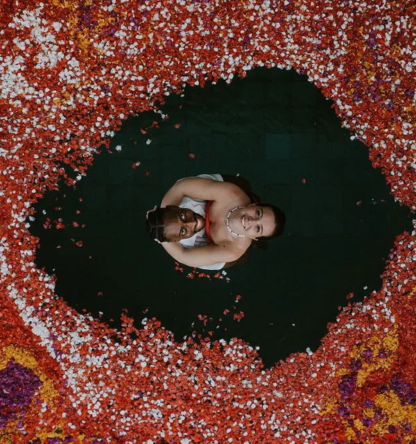 Vue aérienne d'un couple souriant enlacé dans une piscine noire entourée de pétales de fleurs rouges, blanches, jaunes et violettes.