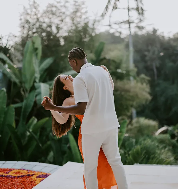 Un couple danse joyeusement au bord d'une piscine entourée de végétation luxuriante, la femme porte une robe orange et l'homme porte une tenue blanche.