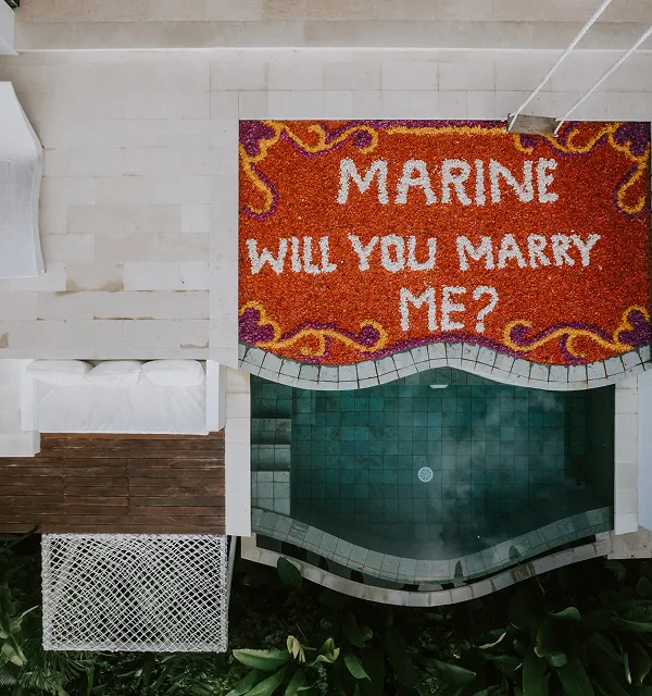 Proposition de mariage vue du dessus avec un tapis de fleurs rouges, blanches, et violettes formant le texte "Marine will you marry me?" au bord d'une piscine bleu-vert.