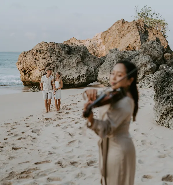 Un couple marchant main dans la main sur une plage de sable près de gros rochers avec une violoniste jouant au premier plan.