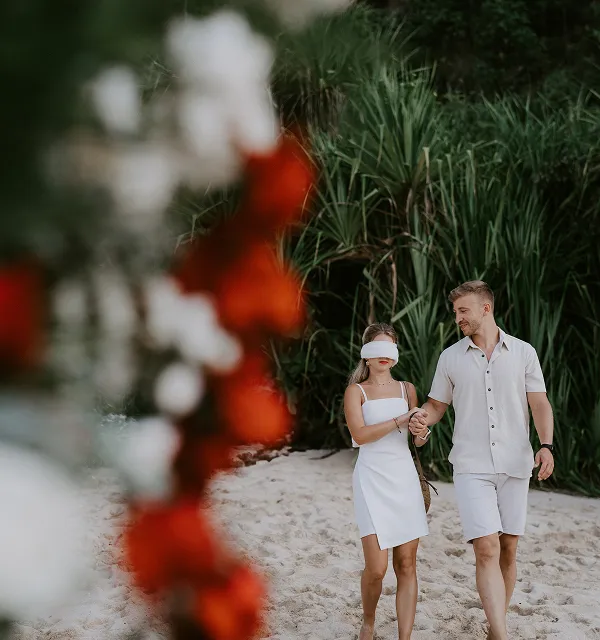 Couple marchant main dans la main sur une plage sableuse avec une femme portant un bandeau blanc sur les yeux, entourée de végétation verte et de fleurs rouges et blanches floues en premier plan.
