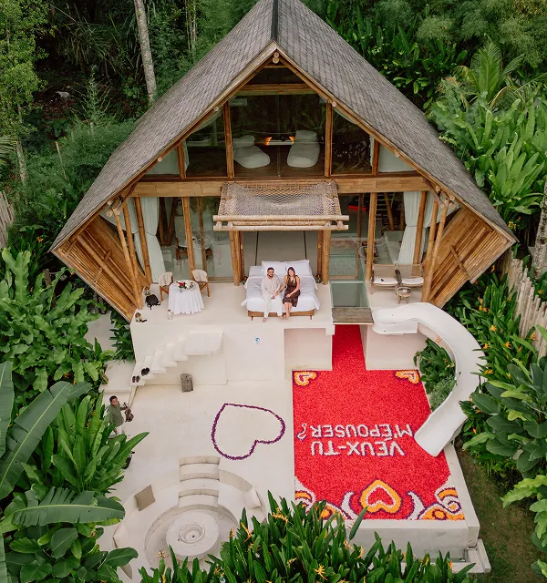 Cabane en bambou avec piscine et tapis de pétales rouges formant un cœur et le texte « Veux-tu m'épouser ? » devant un couple assis, entourée de végétation luxuriante.