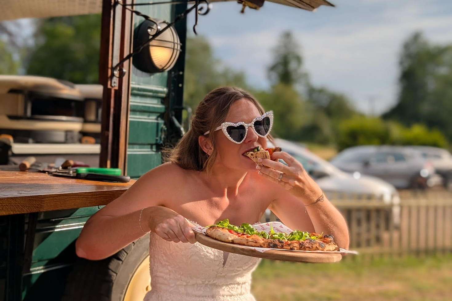 Bride enjoying a slice of freshly baked Frenchie’s Pizza beside the horsebox at an outdoor wedding.