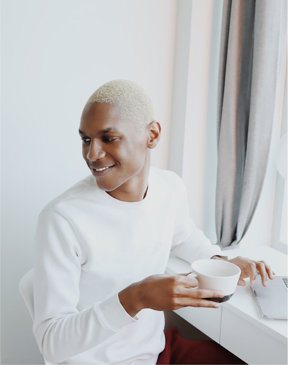 Young person with short blonde hair in a white sweater holding a coffee cup and smiling while sitting at a desk with a laptop.