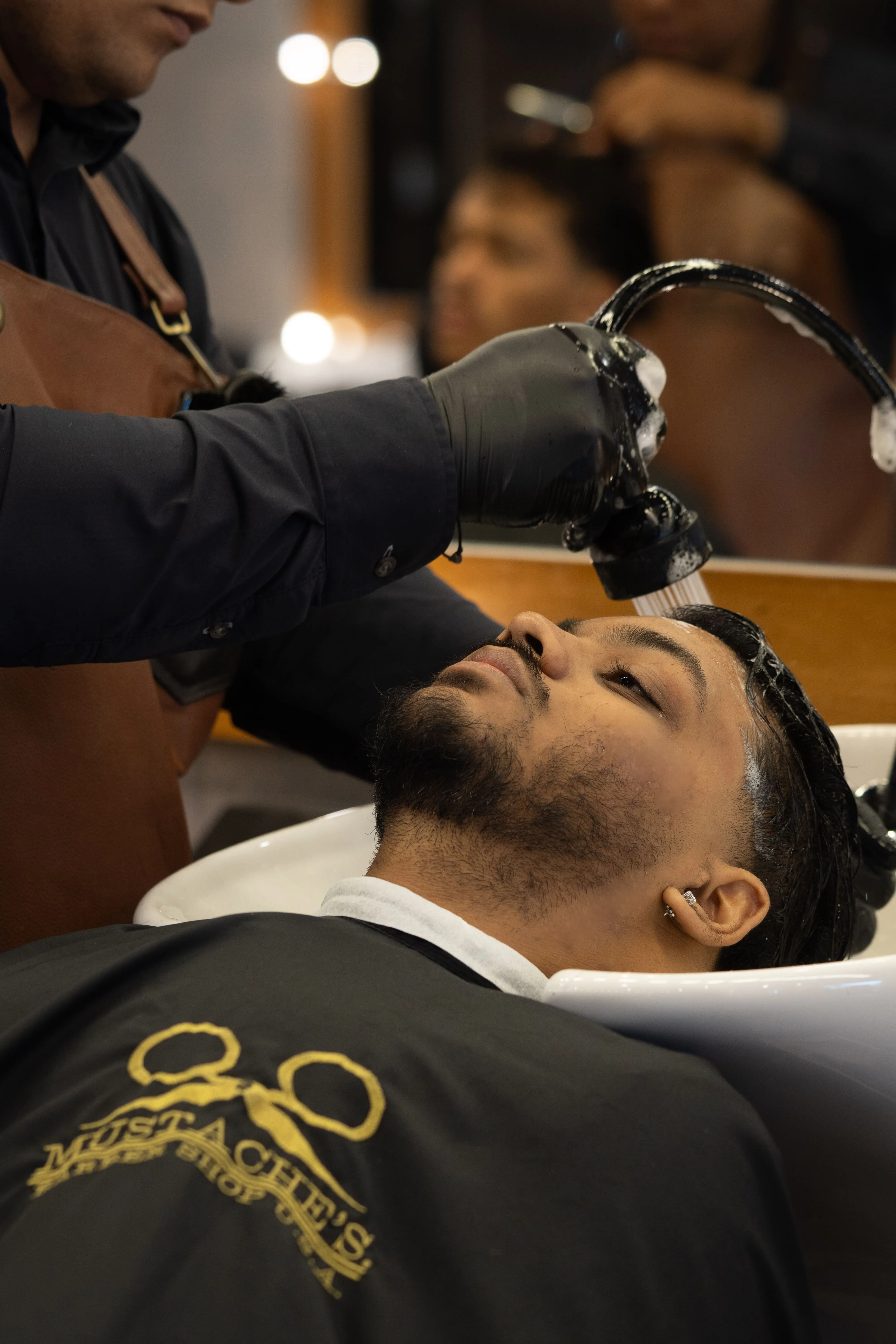 A client lies back as a gloved barber rinses his hair at a wash basin in the barbershop.
