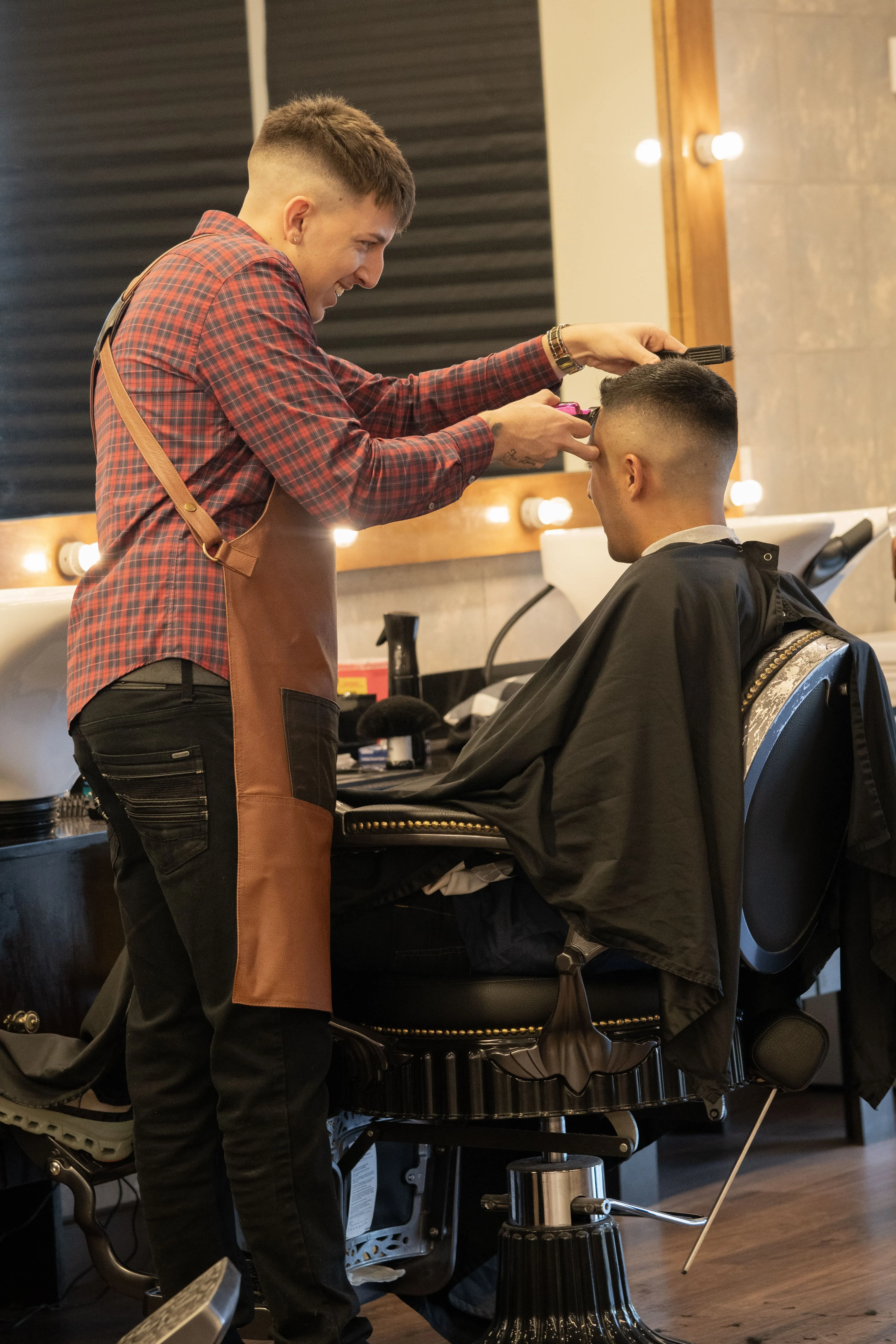 A smiling barber in a plaid shirt and apron gives a client a precise haircut in a modern barbershop.