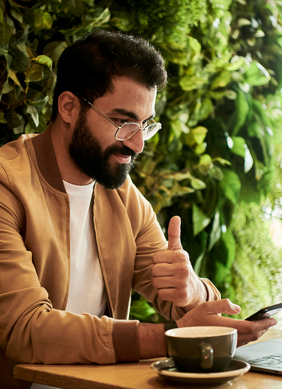 Hombre con gafas y chaqueta marrón haciendo un gesto de aprobación con el pulgar mientras mira un teléfono móvil frente a una taza de café.