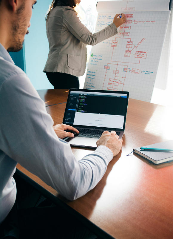 Dos personas en una reunión de trabajo: una mujer escribe en un rotafolio con diagramas y un hombre usa una laptop con código visible en pantalla.