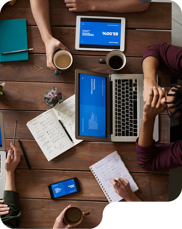 Personas trabajando juntas en una mesa de madera con laptop, tabletas, teléfono inteligente, cuadernos con notas y tazas de café.