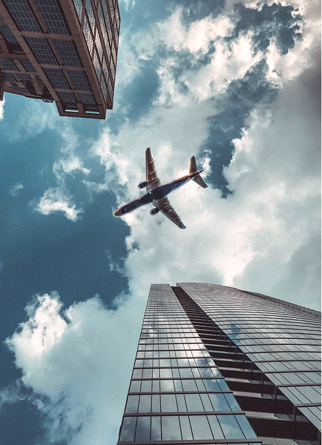 Avión volando entre edificios altos bajo un cielo parcialmente nublado.