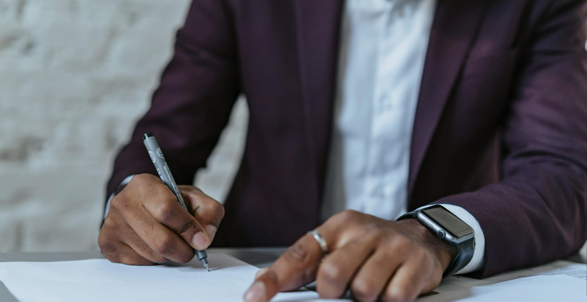 Persona de piel oscura con chaqueta morada y reloj inteligente escribiendo en una hoja blanca con un bolígrafo gris.