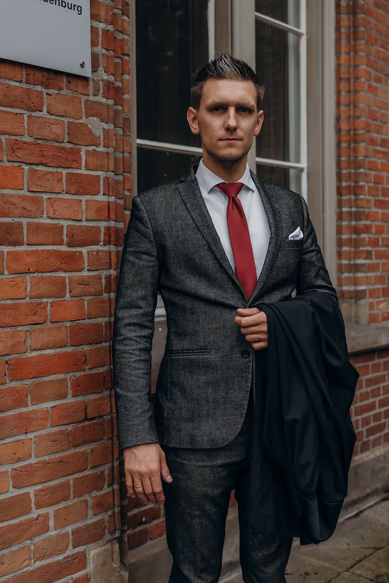Mann im grauen Anzug mit roter Krawatte steht vor einem roten Backsteingebäude und hält einen schwarzen Mantel.