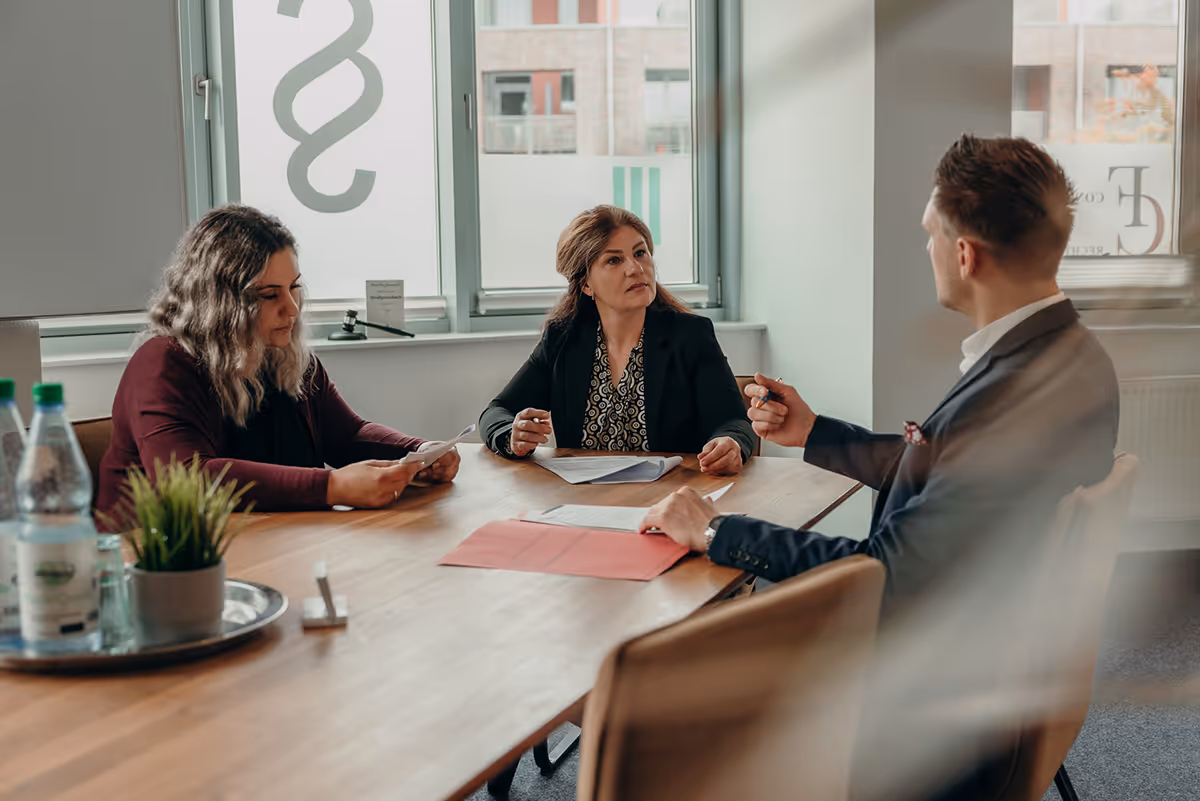 Drei Personen führen ein Gespräch bei einem Besprechungstisch in einem Büro mit großen Fenstern.