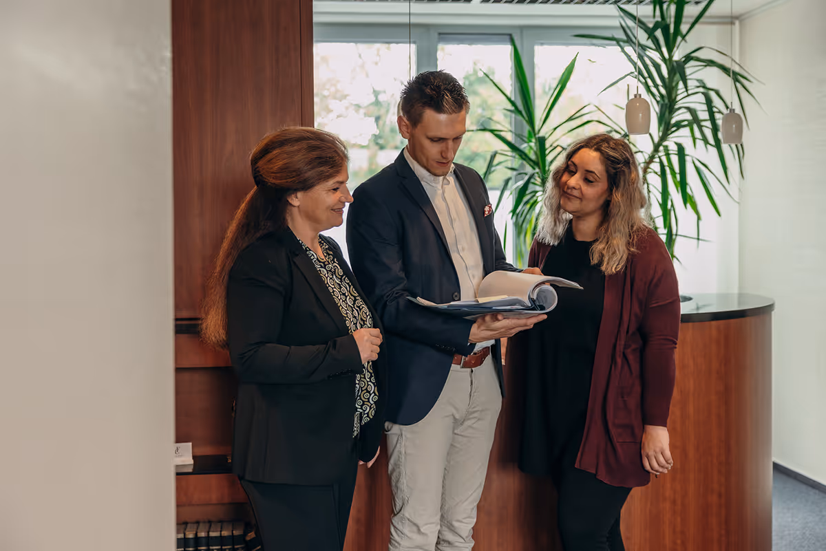 Drei Personen im Büro stehen zusammen und betrachten gemeinsam ein großes geöffnetes Buch.