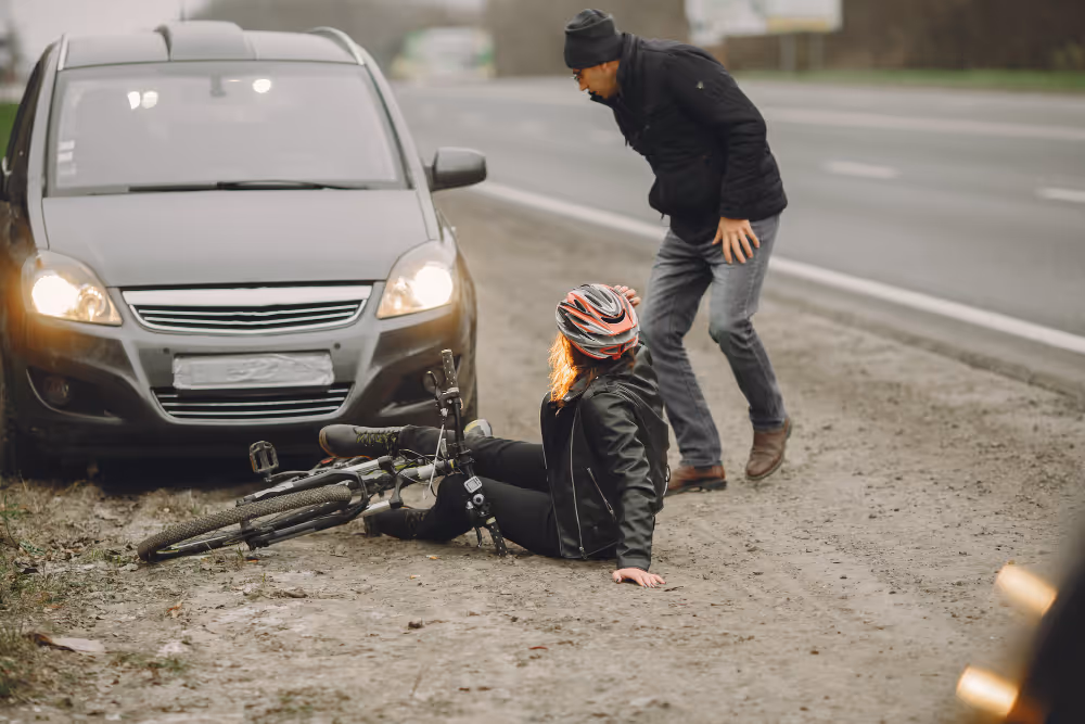 Radfahrerin mit Helm sitzt neben ihrem umgestürzten Fahrrad am Straßenrand, während ein Mann sich sorgenvoll zu ihr beugt.