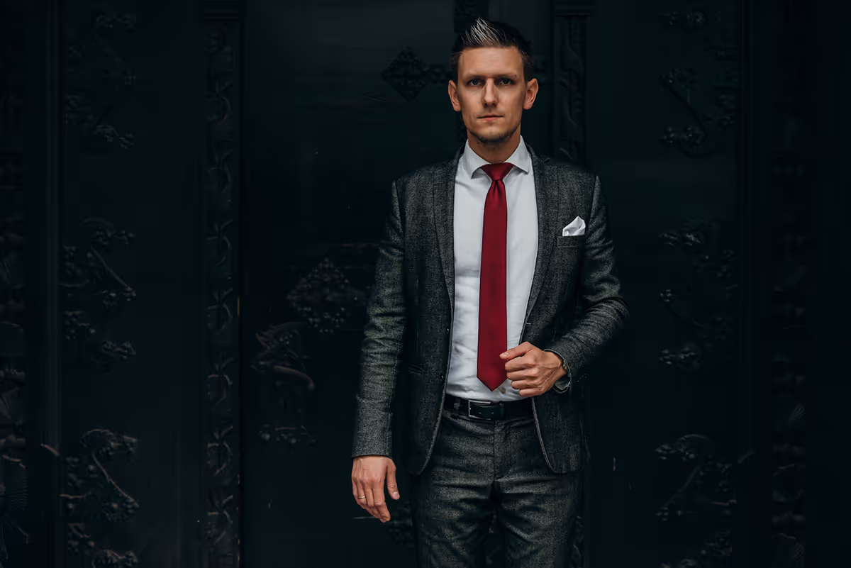 Mann im grauen Anzug mit roter Krawatte und weißem Hemd steht vor dunklem Hintergrund.