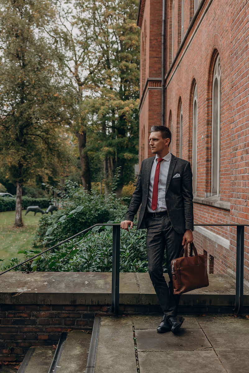 Geschäftsmann in dunkelgrauem Anzug mit roter Krawatte und brauner Aktentasche lehnt an einem Geländer vor einem roten Ziegelgebäude.
