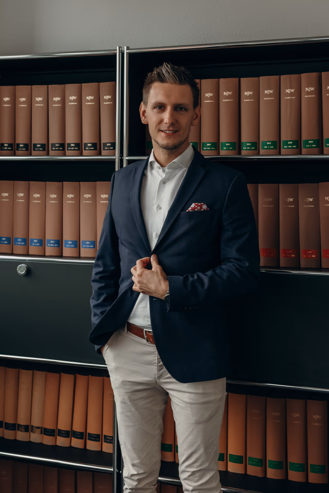 Junger Mann in Anzugjacke, weißem Hemd und heller Hose steht vor einem Bücherregal mit braunen Aktenordnern.