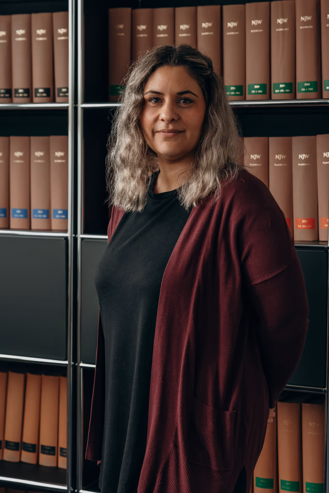 Frau mit schulterlangem, gewelltem Haar trägt schwarzen Pullover und roten Cardigan vor einem Bücherregal mit braunen Rechtsfachbüchern.