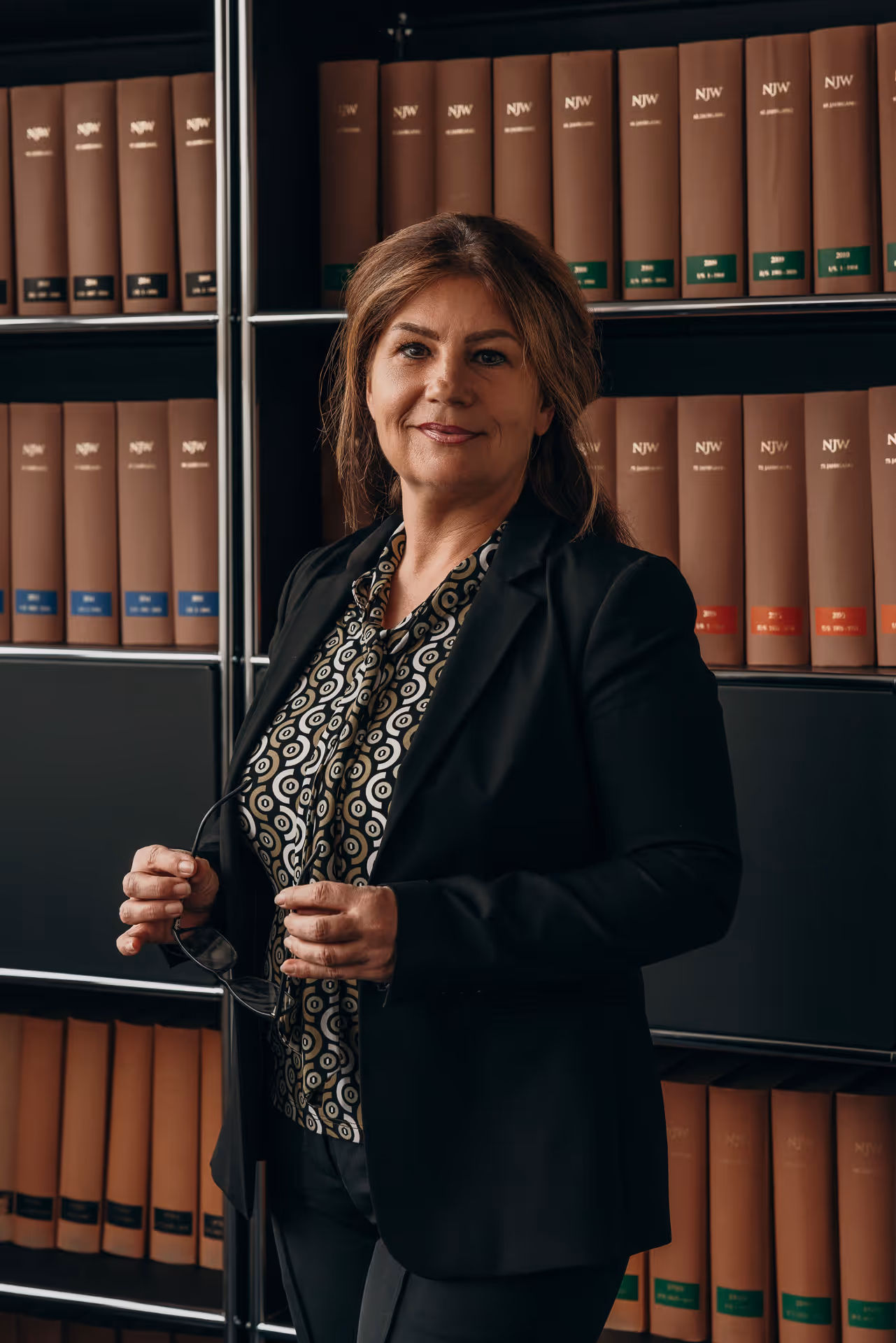 Frau in schwarzem Blazer und gemustertem Hemd steht vor einem Regal mit braunen Gesetzesbüchern und hält eine Brille in den Händen.