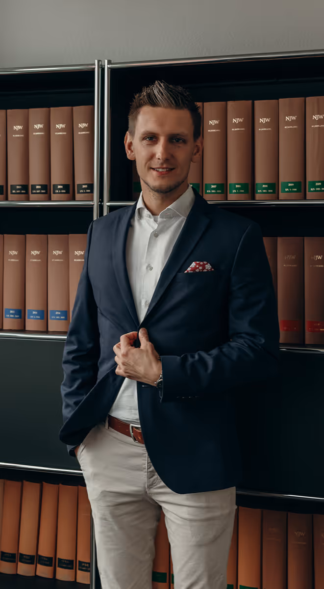 Junger Mann in dunkelblauem Sakko und weißem Hemd steht vor einem Regal mit ordentlichen braunen Aktenordnern.