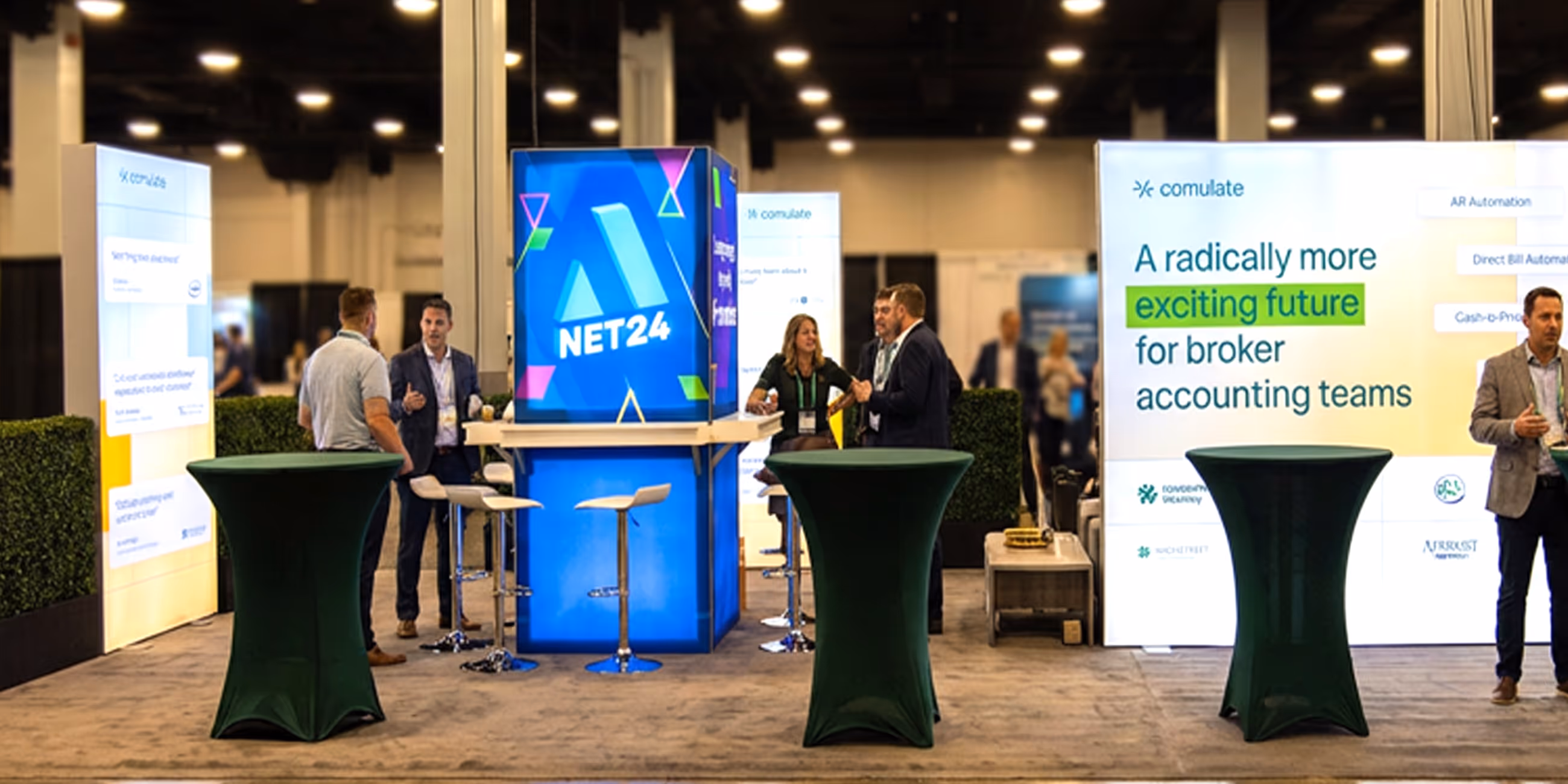 Trade show booth with a bright blue NET24 display, green-covered cocktail tables, and people conversing.