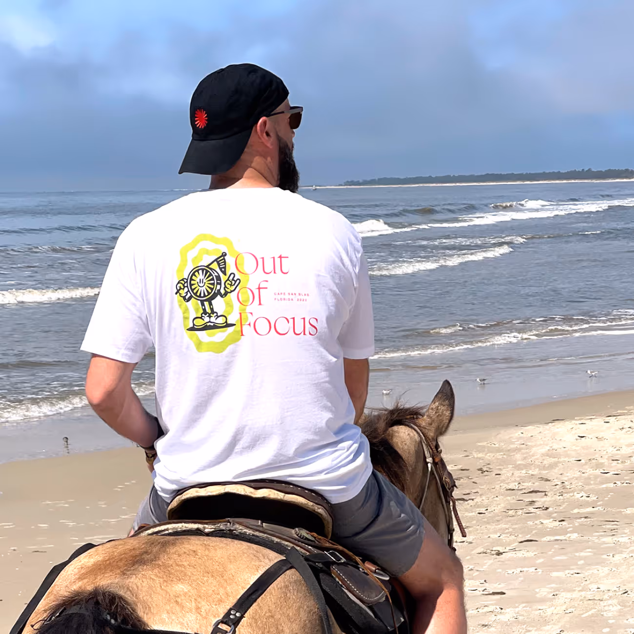 Man wearing a white 'Out of Focus' t-shirt riding a horse on a sandy beach with ocean waves in the background.