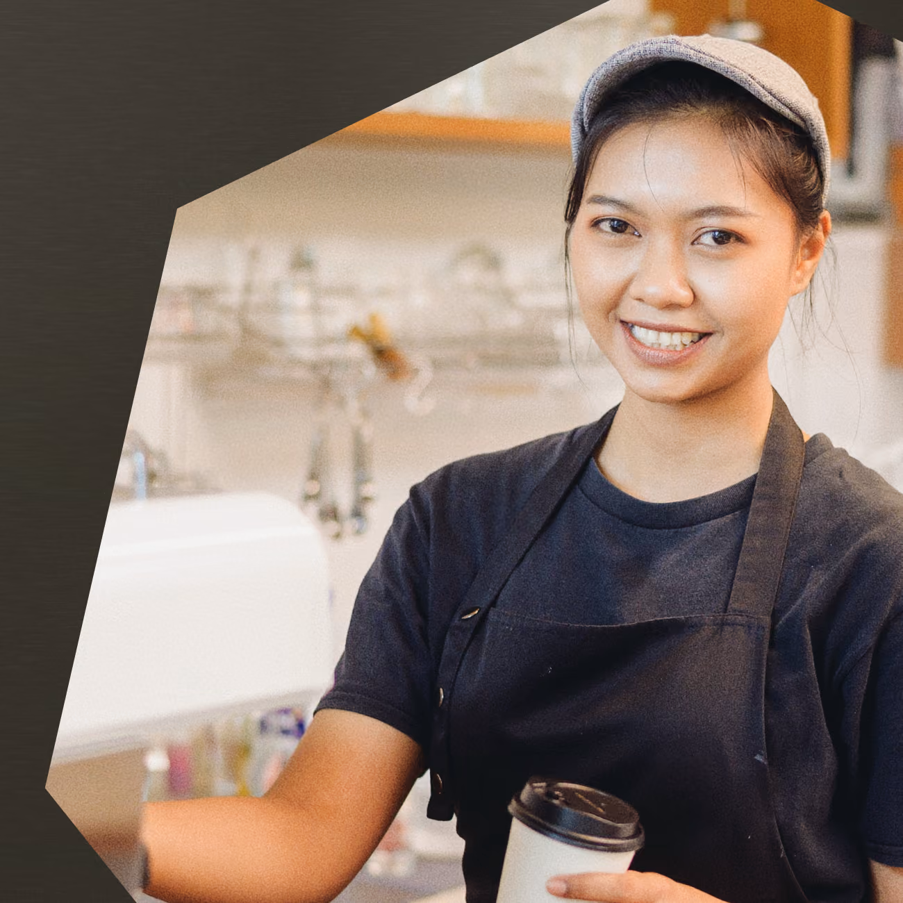 Smiling woman wearing a gray cap and black apron holding a takeaway cup in a coffee shop.