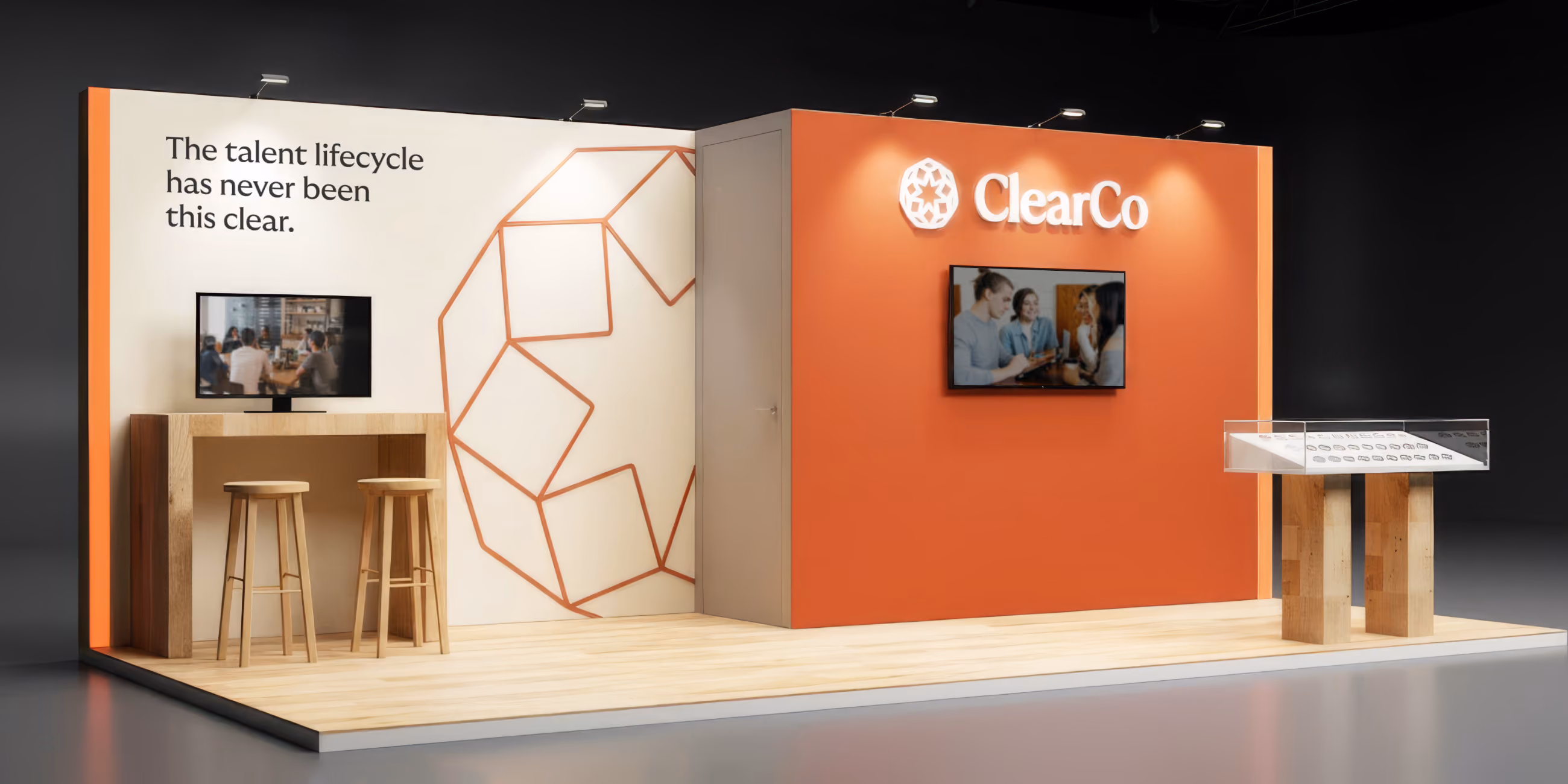 ClearCo trade show booth with orange and white walls, one side showing the text 'The talent lifecycle has never been this clear,' two stools by a wooden counter with a TV, and the other side with the ClearCo logo above a mounted screen, plus a small table display on the right.