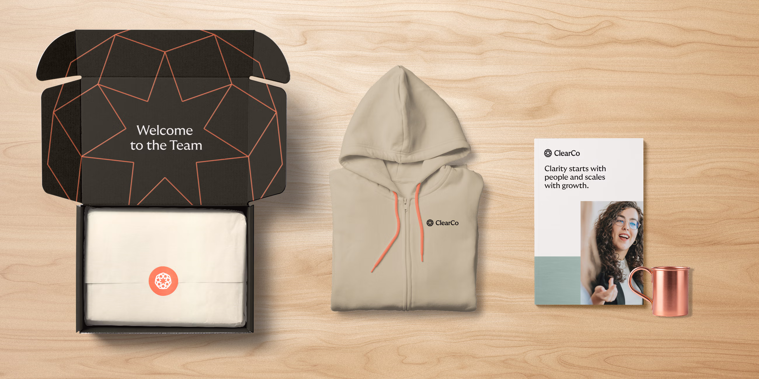 Welcome kit including an open black box with 'Welcome to the Team' text inside and a folded white item inside, a beige ClearCo-branded zip-up hoodie, a ClearCo pamphlet with text 'Clarity starts with people and scales with growth,' and a copper cup, arranged on a light wood surface.